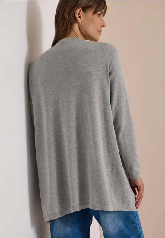 Frau von hinten in grauem, locker fließendem Strickcardigan mit blauen Jeans, lehnt an hellem Hintergrund.