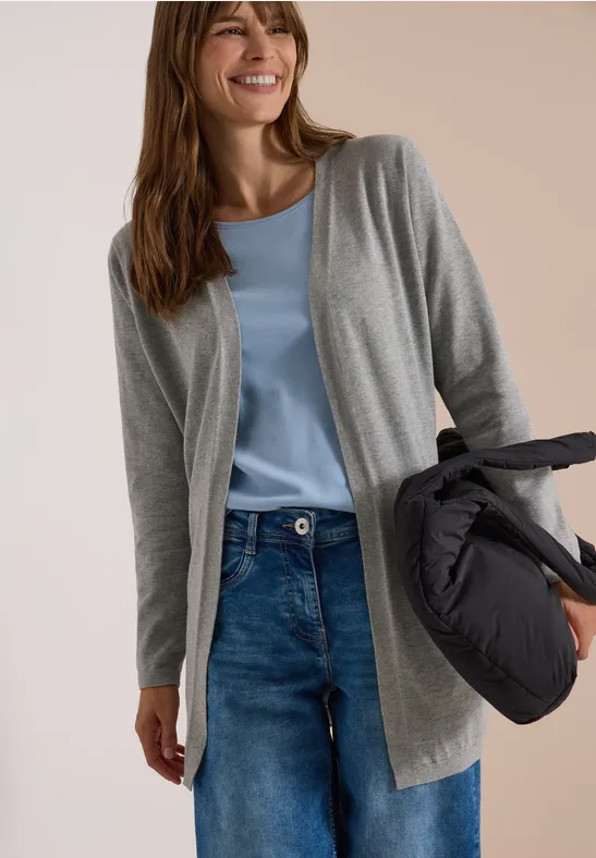 Eine lächelnde Frau trägt graue Strickjacke, hellblaues Top, Jeans und hält eine Daunenjacke.