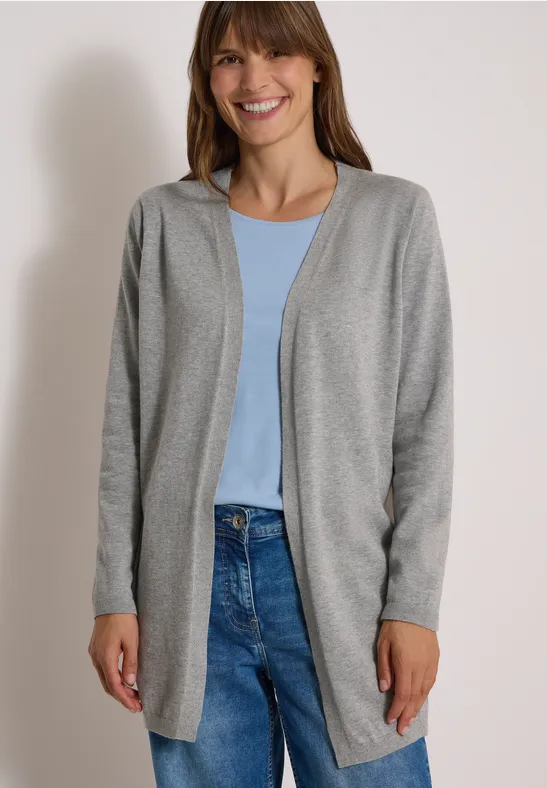 Frau im grauen Cardigan, blauem Shirt und Jeans lächelt vor neutralem Hintergrund.