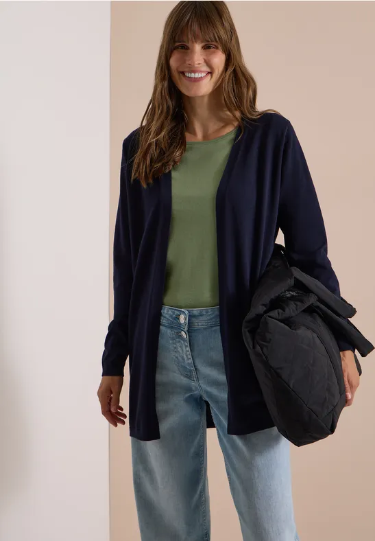 Eine lächelnde Frau trägt grüne Bluse, dunkelblaue Jacke, Jeans und hält eine schwarze Tasche.