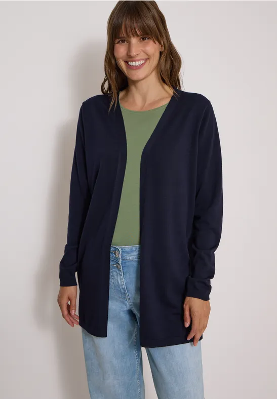 Frau lächelt, trägt dunkelblaue Strickjacke über grünem Shirt und Jeans.