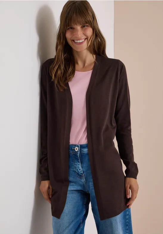 Frau lehnt lächelnd an Wand, trägt rosa Shirt, offene braune Strickjacke und blaue Jeans.