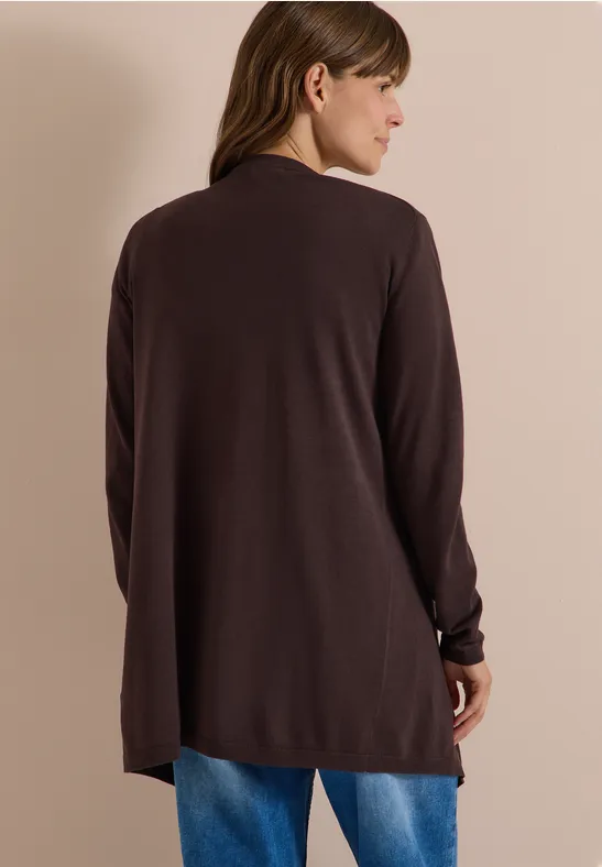 Frau von hinten in langem, dunklem Cardigan und blauer Jeans vor neutralem Hintergrund.