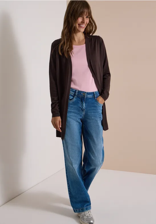 Lächelnde Frau in blauen Jeans, rosa Top und schwarzer Strickjacke vor neutralem Hintergrund.