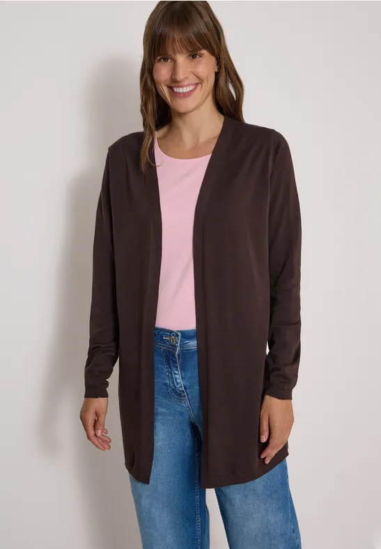 Eine lächelnde Frau trägt eine dunkelbraune offene Strickjacke, ein rosafarbenes T-Shirt und blaue Jeans.