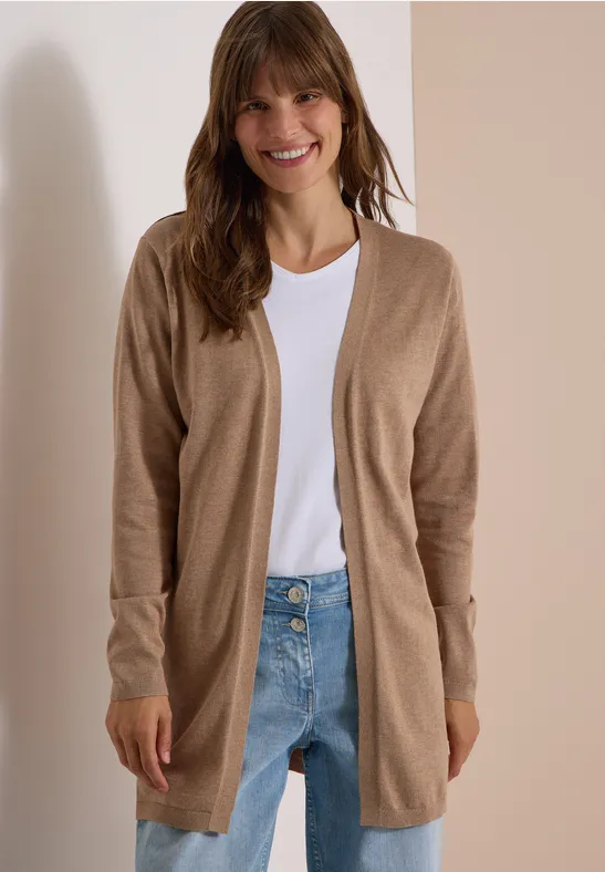 Eine lächelnde Frau in beigefarbener Strickjacke, weißem Shirt und Jeans vor hellem Hintergrund.