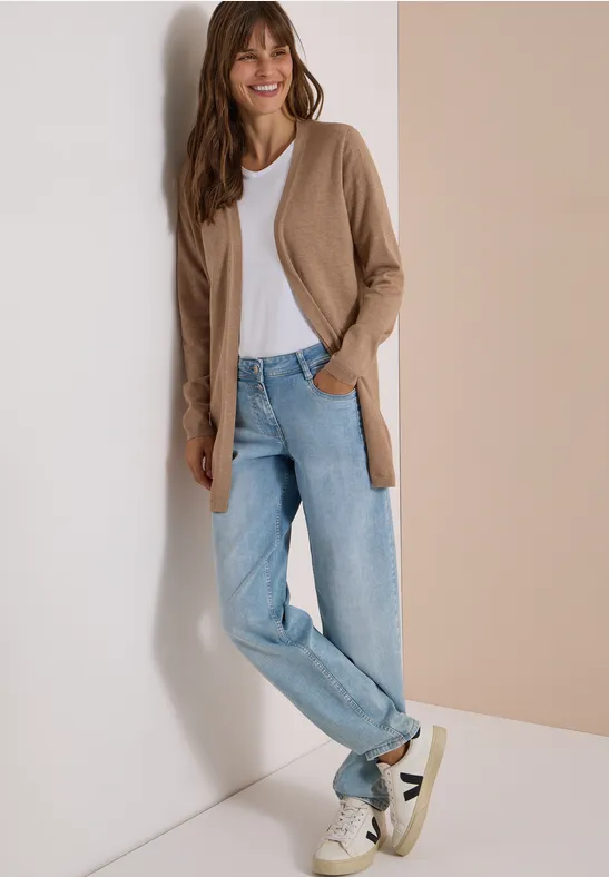 Eine Frau lehnt an einer Wand, trägt hellblaue Jeans, weißes T-Shirt, braunen Cardigan und weiße Sneakers.