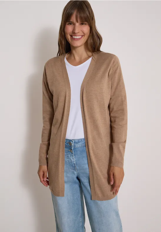 Frau in beige Cardigan, weißem T-Shirt und Jeans lächelt vor weißer Wand.