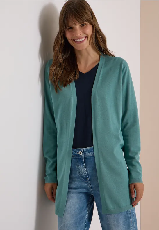 Junge Frau lehnt lächelnd an einer Wand, trägt grünen Cardigan, dunkelblauen Shirt und Jeans.