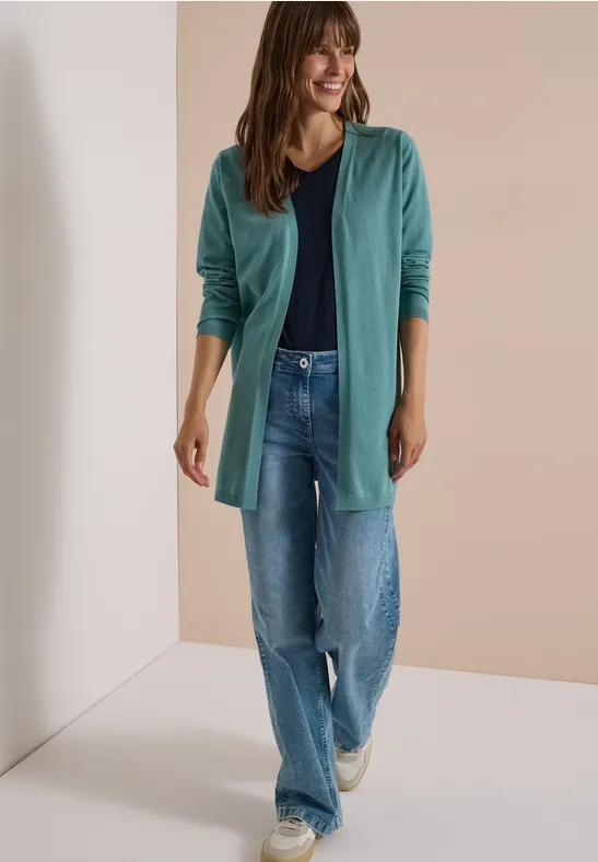 Eine Frau posiert lächelnd in türkisfarbenem Cardigan, dunklem Top, Jeans und weißen Sneakern.
