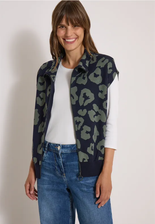 Frau in Leopardenmuster-Strickjacke, weißem Shirt und Jeans, lächelt vor neutralem Hintergrund.
