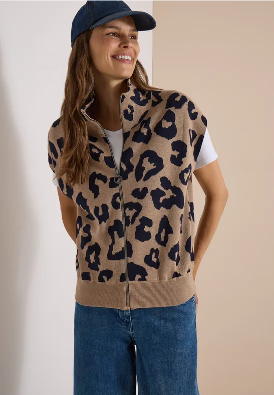 Frau mit beigem Strick-Gilet im Leopardenmuster, weißem Shirt, blauen Jeans und navy Cap lächelnd.