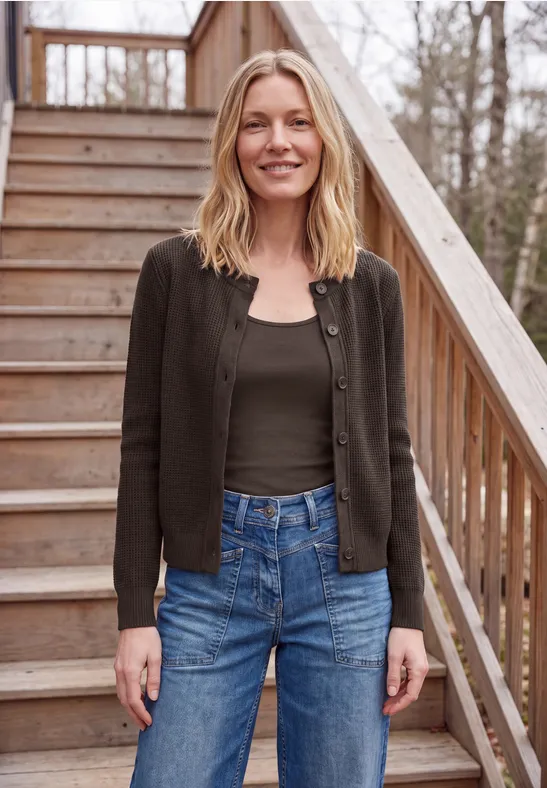Frau steht lächelnd auf einer Holztreppe im Freien, trägt schwarze Jacke, grünes Top und blaue Jeans.
