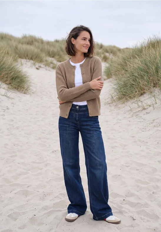 Eine Frau in Jeans und Strickjacke steht entspannt zwischen Dünengräsern am Strand.