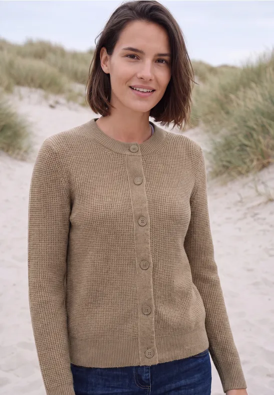 Frau mit kurzem braunem Haar in beiger Strickjacke steht entspannt am Dünenstrand.