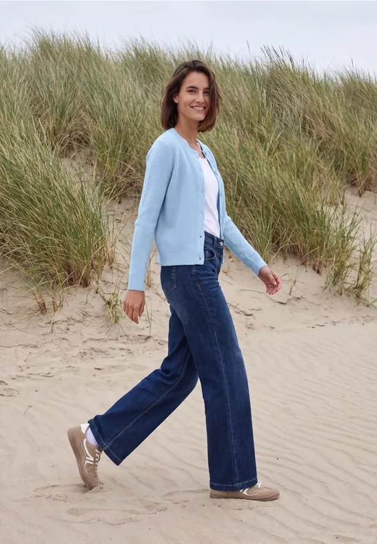 Eine Frau in Jeans, weißem Shirt und hellblauer Strickjacke spaziert über Dünen am Strand.