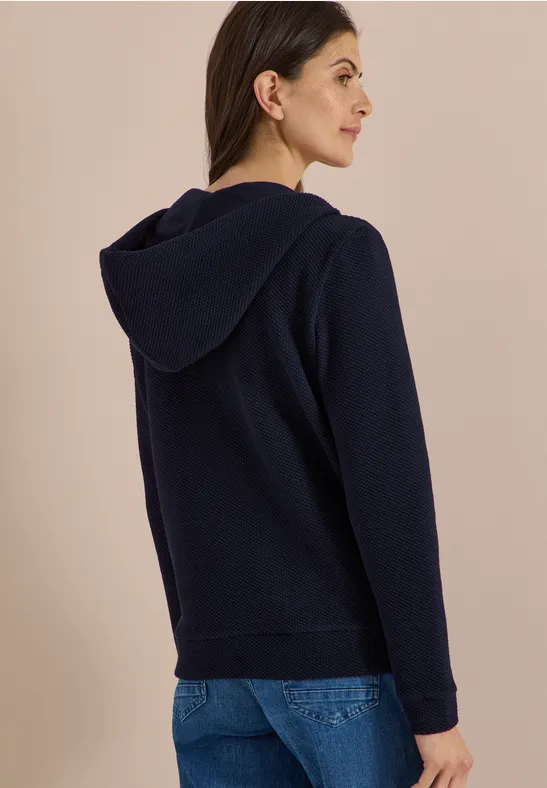 Frau von hinten in dunkelblauer Kapuzenjacke und Jeans vor beiger Wand.