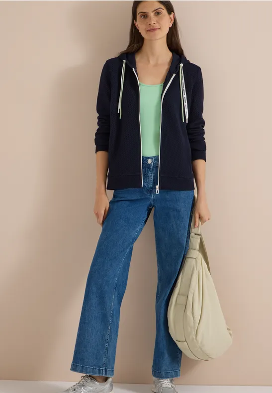 Eine junge Frau in navyblauem Hoodie, mintgrünem Top, weiten Jeans und großer Stofftasche vor beigem Hintergrund.