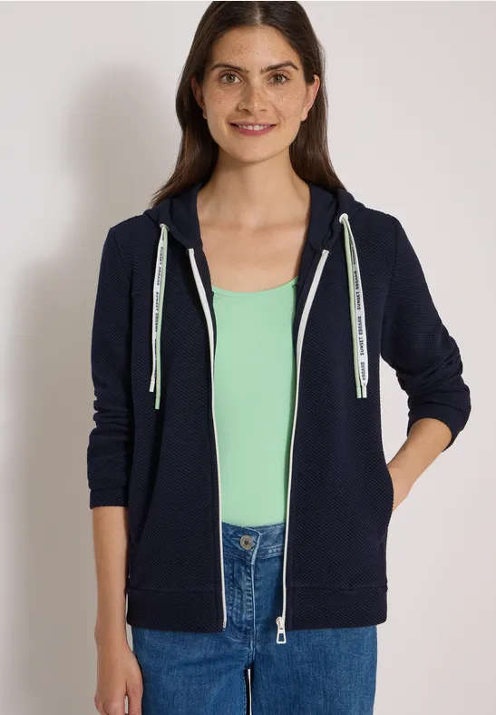Eine lächelnde Frau in dunkelblauem Kapuzen-Sweatshirt, grünem Top und Jeans.