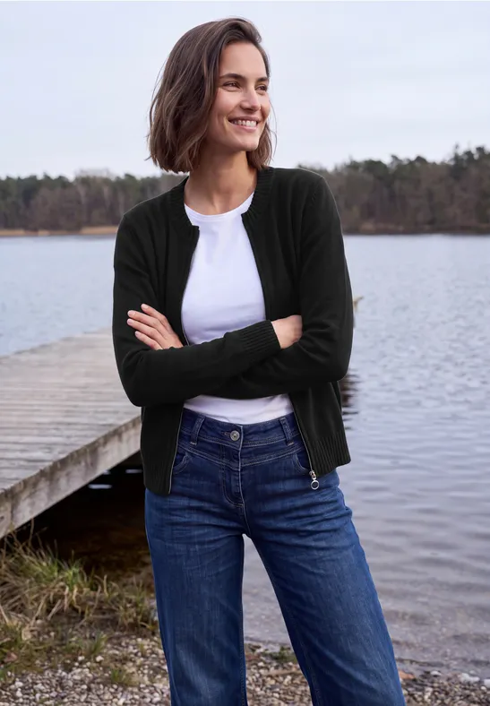 Vrouw in jeans, wit shirt en donker vest, glimlachend met armen over elkaar, staand op een steiger bij een meer.