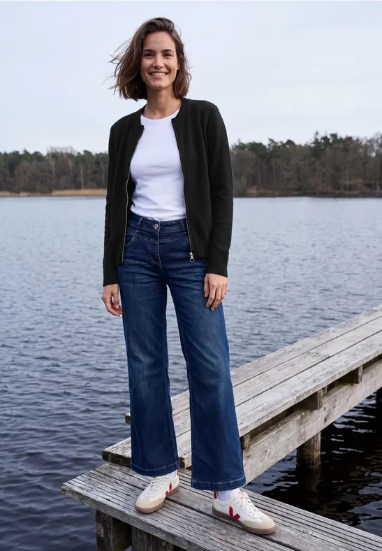 Vrouw staat lachend op steiger bij meer, draagt wit shirt, donkere jas, wijde jeans en witte instapschoenen.