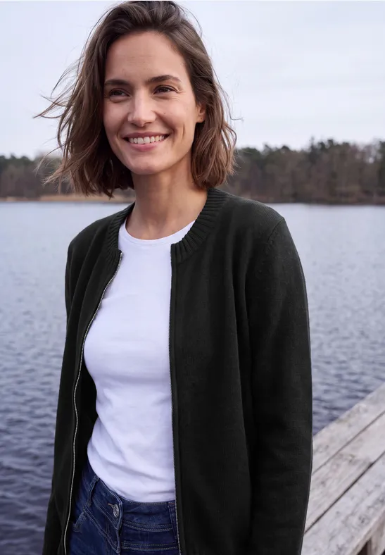 Een glimlachende vrouw in wit t-shirt en donkergroene cardigan staat op een steiger bij het meer.