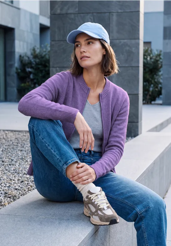 Junge Frau sitzt in lila Strickjacke, Jeans und Baseballcap auf moderner Bank im urbanen Umfeld.