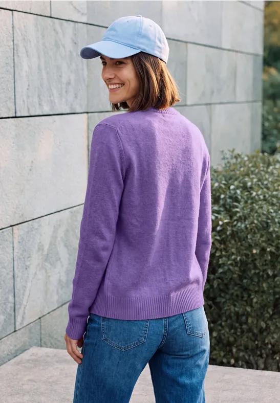 Frau in lila Pullover, Jeans und weißer Mütze lächelt seitlich vor einer Steinwand.