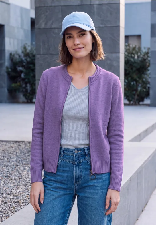 Frau mit lila Cardigan, grauem Top, Jeans und blauer Schirmmütze steht im Freien.