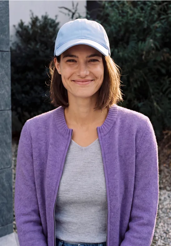 Eine lächelnde Frau in fliederfarbener Strickjacke, grauem Shirt und hellblauer Baseballkappe steht draußen.