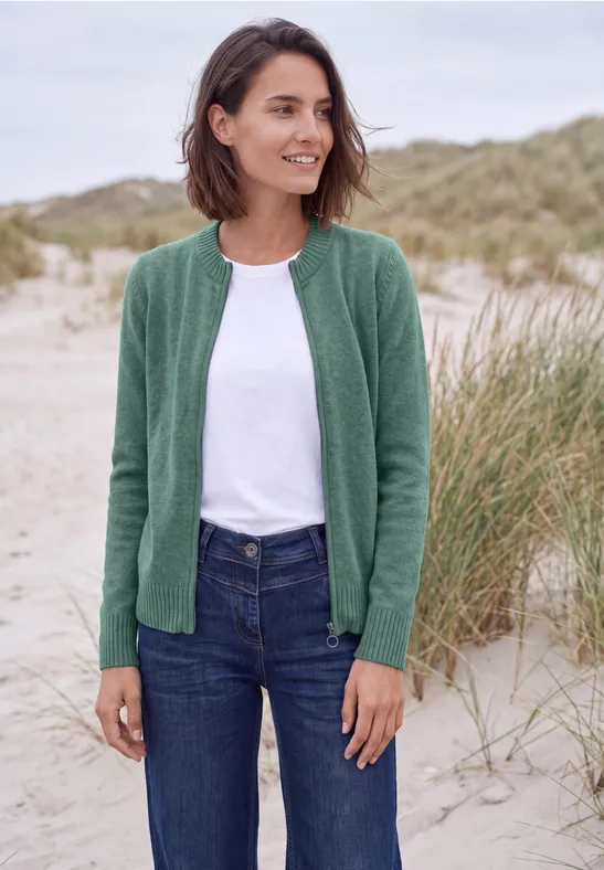 Eine Frau in grüner Jacke, weißem T-Shirt und Jeans steht lächelnd am Strand vor Dünen.
