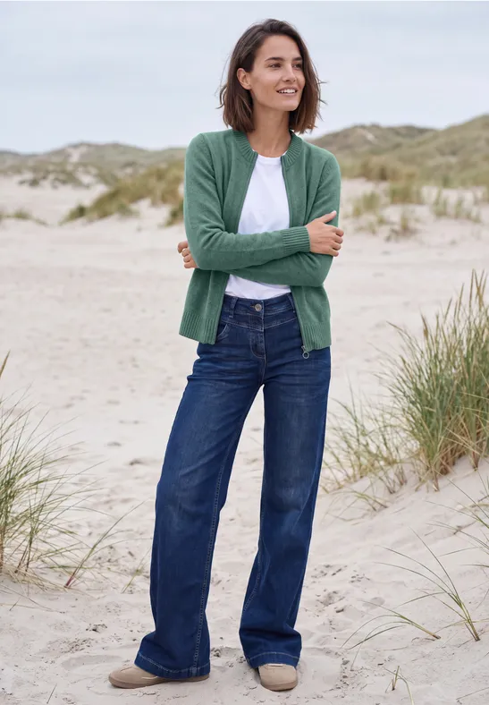 Eine lächelnde Frau steht am Sandstrand vor Dünen, trägt grüne Strickjacke, weißes Shirt und weite Jeans.