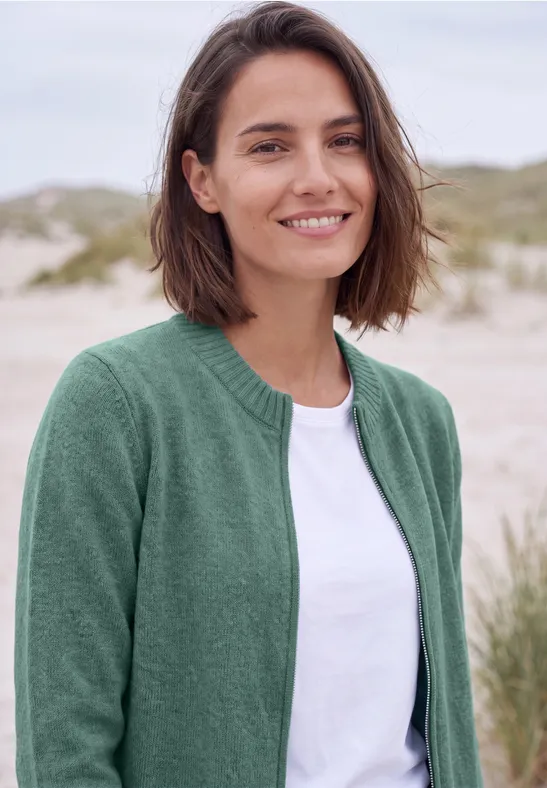 Eine lächelnde junge Frau in grüner Strickjacke und weißem Shirt steht am Strand.