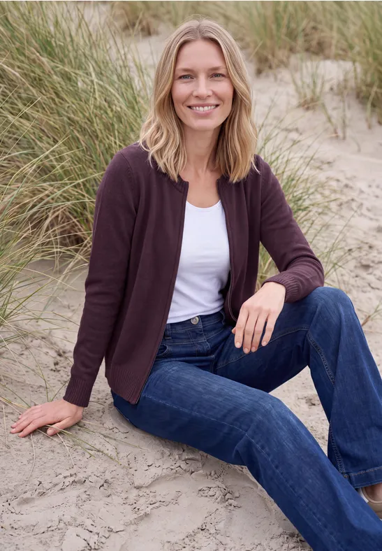 Frau sitzt entspannt im Stranddünensand, trägt Jeans, weißes Shirt und Strickjacke, lächelt.