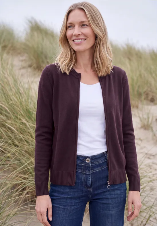 Frau im lila Cardigan und Jeans steht lächelnd in Dünen mit Strandgras.