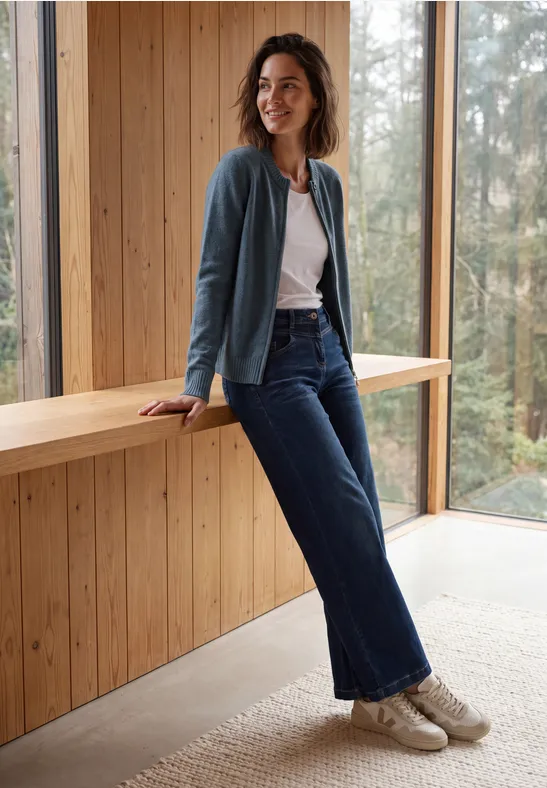 Vrouw in jeans en vest leunt ontspannen op een houten bank in een moderne, lichtdoorlatende ruimte.