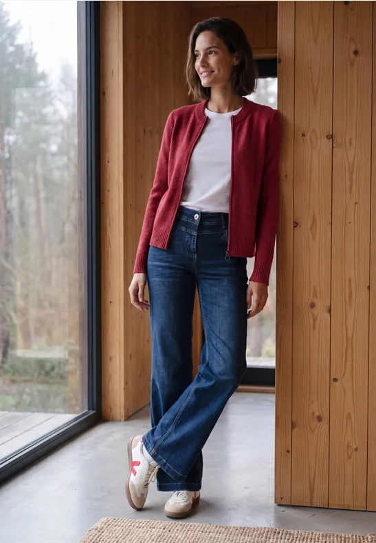 Frau in roter Strickjacke, weißem Shirt und Jeans lehnt entspannt am Fenster in modernem Holzhaus.