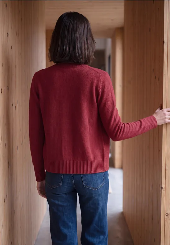 Eine Person steht mit dem Rücken zur Kamera in einem Holzflur, trägt roten Pullover und blaue Jeans.