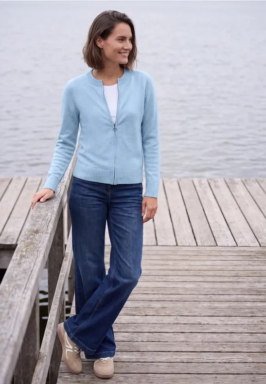 Frau mit blauem Cardigan und Jeans steht lächelnd auf einem Steg am See.