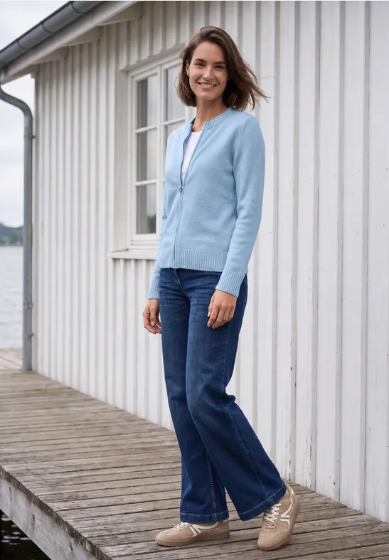 Frau lächelt auf Holzsteg am Wasser neben weißem Haus, trägt hellblaue Jacke, Jeans und bequeme Schuhe.