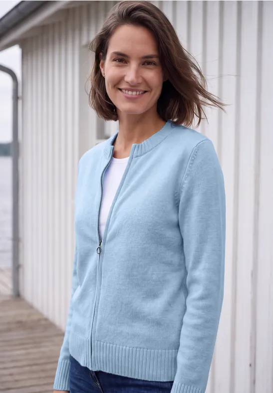 Lächelnde Frau in hellblauer Strickjacke vor weißem Holzhäuschen.