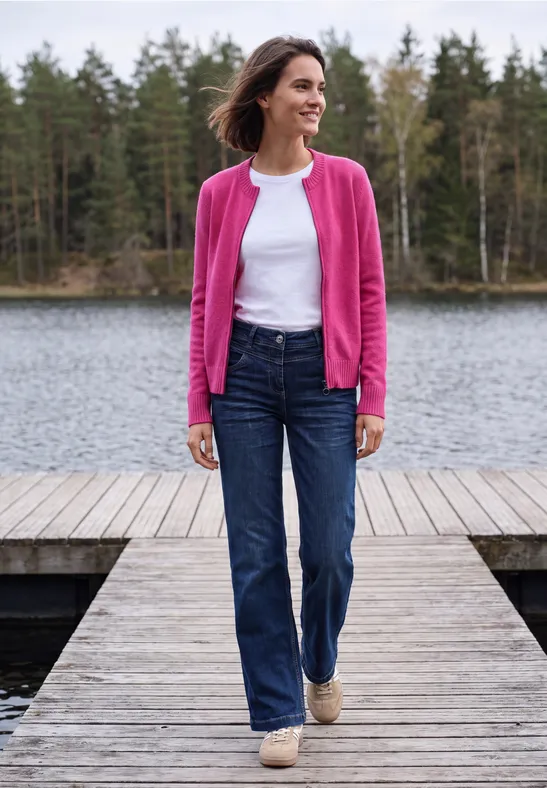 Frau in Jeans, weißem Shirt und pinkem Cardigan steht auf einem Holzsteg am See, umgeben von Wald.