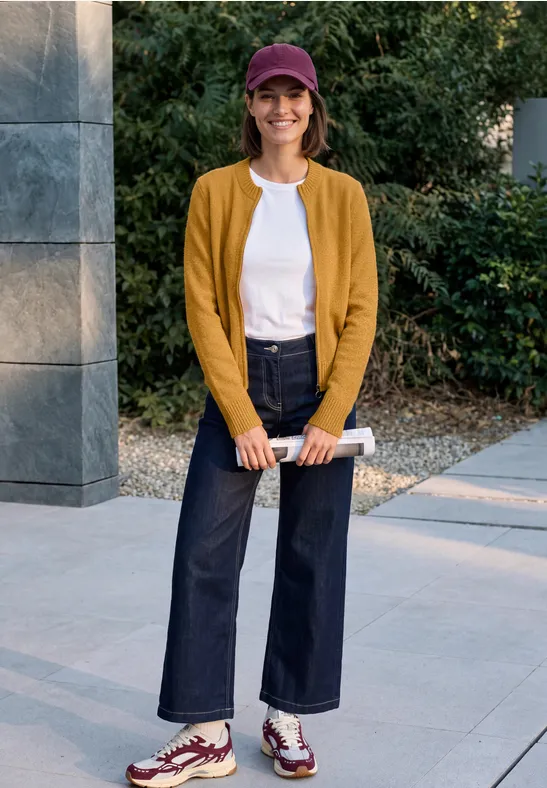 Person mit roter Kappe, senfgelbem Cardigan, weißem Shirt und weiten Jeans steht draußen und hält ein gerolltes Magazin.