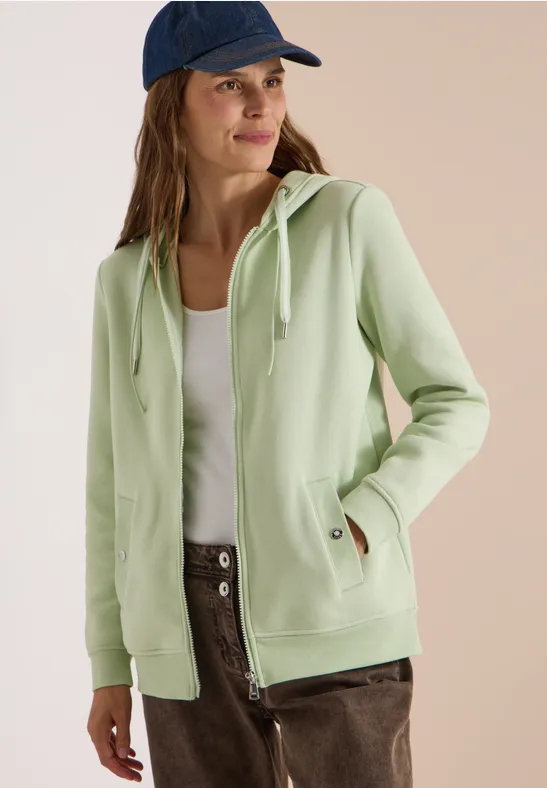Frau in pastellgrüner Kapuzenjacke, weißem Top, brauner Hose und blauer Mütze lächelt seitlich.
