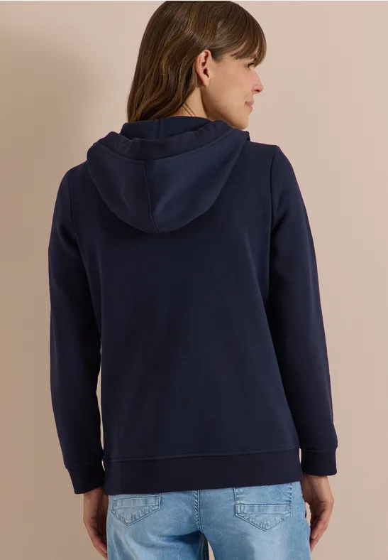 Frau von hinten in dunkelblauer Kapuzenjacke und hellen Jeans vor neutralem Hintergrund.