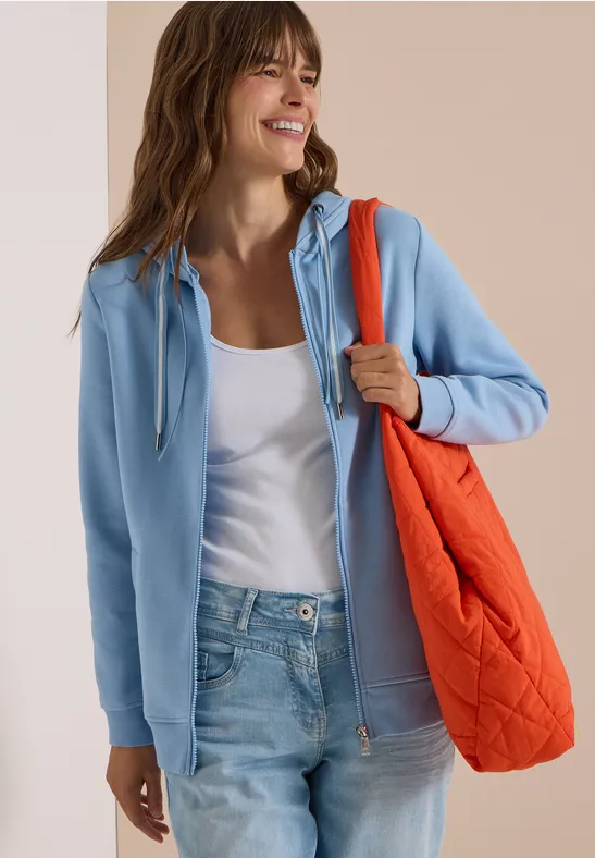 Frau in hellblauer Kapuzenjacke, weißem Top, Jeans und orangefarbener, gesteppter Tasche.