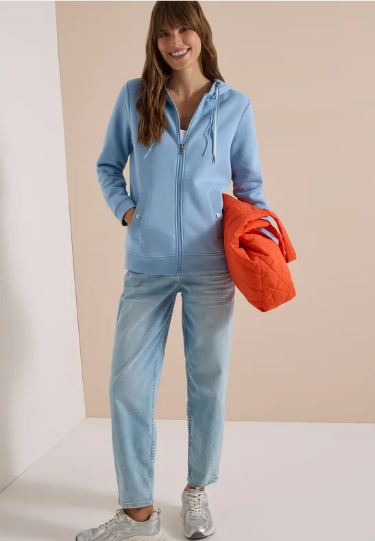 Frau in hellblauer Kapuzenjacke, Jeans und grauen Sneakern, mit orangefarbener Stepp-Tasche.
