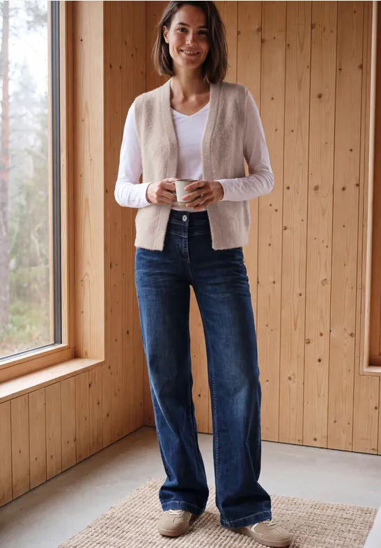 Eine Frau steht in einem hellen Holzraum am Fenster, trägt Jeans, Cardigan und lächelt.