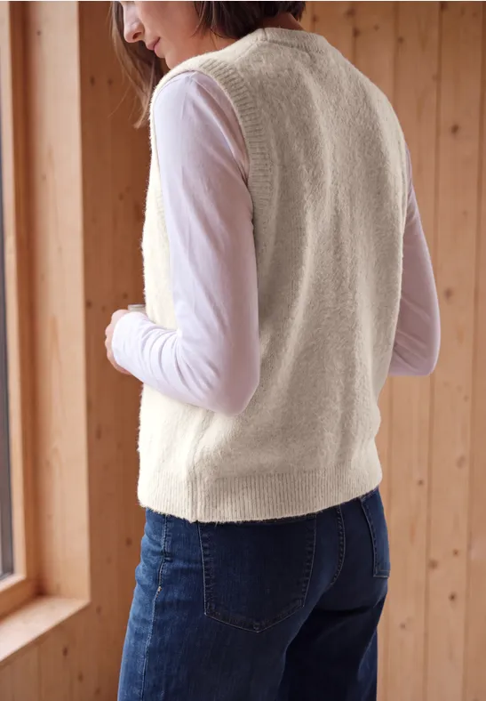 Frau von hinten in beige Fleece-Weste und weißem Langarmshirt vor einem Fenster in Holzraum.