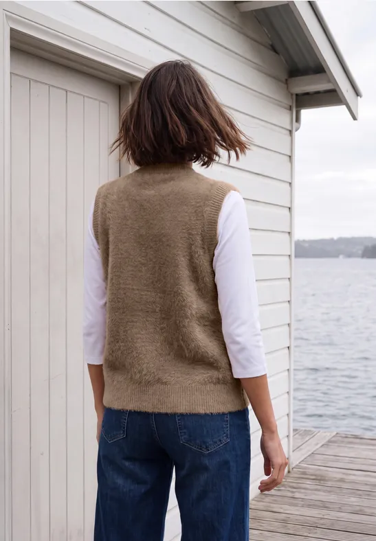 Eine Frau steht in Weste und Jeans auf einem Steg vor einem Bootshaus und blickt aufs Wasser.